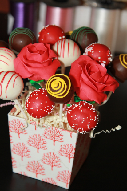 An array of gourmet cake mix cake pops adorned with various decorations, including red with white sprinkles, white with red drizzles, and some designed to resemble red roses and yellow swirls, presented in a white box with a red coral tree pattern, set against a backdrop of ribbon rolls.